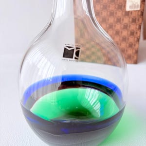 May include: A clear glass vase with a rounded body and a narrow neck. The base features horizontal bands of deep blue and vibrant green glass. A square black and white label with the text "MADE IN ITALY" and "carlo moretti" is on the front.