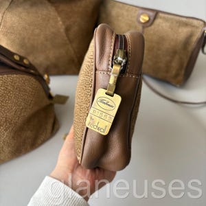 May include: A close-up of a brown leather clutch with a textured surface and a gold zipper. The clutch has a rectangular shape and a tag that reads "Borbonese by redwall". Other brown bags are in the background.