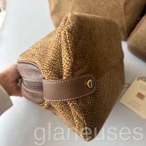 May include: A brown textured cosmetic bag with a brown leather handle and zipper. The bag has a gold-colored clasp and a brown leather accent. The bag is made of a textured material and has a rectangular shape.
