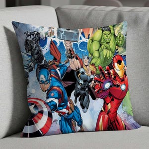 May include: Square throw pillow featuring a comic book style illustration of several Marvel superheroes, including Captain America, Iron Man, Thor, Hulk, and Black Panther. The pillow has a white background and is placed on a gray couch.