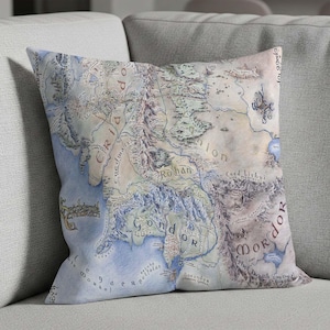 May include: A decorative throw pillow featuring a detailed map of a fantasy world. The map is in shades of blue, beige, and brown, with geographical features and text labels. The pillow is square and rests on a gray sofa.