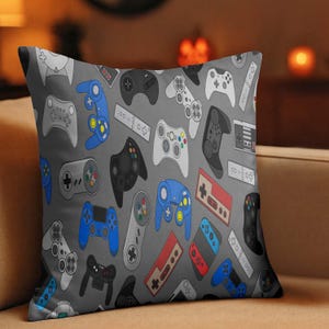 May include: Square throw pillow with a gray background and a pattern of various video game controllers in shades of blue, black, white, and red. The controllers are of different shapes and sizes, creating a playful design.