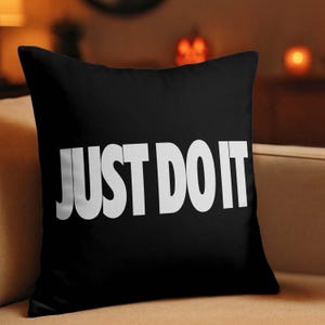 May include: Black square throw pillow with the white text "JUST DO IT" in a bold, sans-serif font. The pillow is on a beige sofa. The background is blurred, with warm lighting.