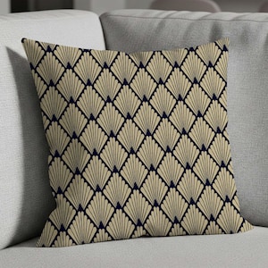 May include: Square decorative pillow with a repeating geometric fan pattern in beige and navy blue. The pillow is placed on a gray sofa. The design is symmetrical and creates a textured appearance.