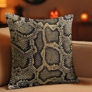 May include: Square decorative pillow with a snakeskin pattern. The pillow features a black and gold design, with a detailed print resembling snake scales. The pillow is placed on a tan sofa, creating a contrast of colors.