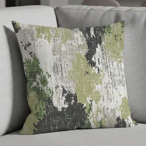 May include: Square decorative pillow with a textured abstract design. The pillow features a pattern of green, gray, black, and white brushstrokes. The pillow is placed on a gray sofa.