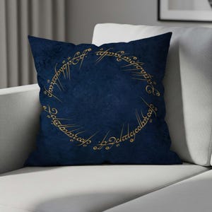 Puede incluir: Un cojín cuadrado azul marino con una inscripción dorada en forma de círculo. El cojín tiene un aspecto texturizado y está colocado sobre un sofá blanco. La inscripción está en una fuente estilizada.