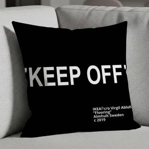 Pode incluir: Almofada quadrada preta com o texto branco "'KEEP OFF'". A almofada também inclui o texto "IKEA® c/o Virgil Abloh 'Flooring' Almhult Sweden c 2019". A almofada está em um sofá cinza.