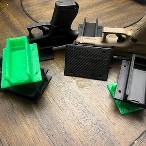 May include: An assortment of gun holsters and firearms. The holsters are in black, green, and carbon fiber patterns. Two black handguns and one tan handgun are also visible.