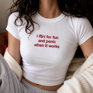 Könnte beinhalten: Weißes Crop-Top mit dem in Rot gestickten Text "I flirt for fun and panic when it works". Das Shirt hat kurze Ärmel und einen Rundhalsausschnitt. Das Model trägt eine graue Jogginghose und eine cremefarbene Strickjacke.