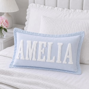 May include: A rectangular light blue throw pillow featuring the name AMELIA in large, white, textured, varsity-style chenille letters centered on the front. The pillow has a matching light blue piped border along all four edges of the fabric.