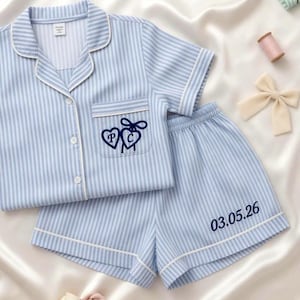 May include: Light blue and white striped pajama set. The short-sleeved top has a breast pocket with embroidered hearts and initials. The shorts have the date "03.05.26" printed on the leg. White piping trims the edges.