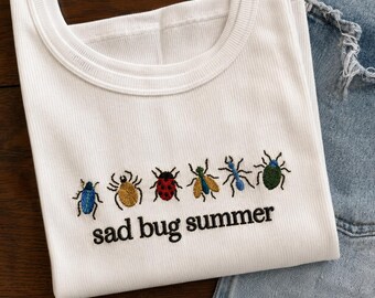 May include: A white ribbed tank top featuring an embroidered row of six colorful insects including a blue beetle, a yellow spider, a red ladybug, a green and yellow fly, a blue ant, and a green beetle, with the text sad bug summer below.
