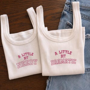 May include: Two white ribbed tank tops featuring pink embroidered text. The left tank top displays the words A LITTLE BIT NEEDY, while the right tank top displays the words A LITTLE BIT DRAMATIC in a collegiate-style font with a pink outline.