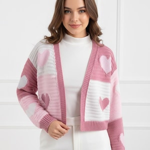 May include: A cropped, open-front cardigan in shades of pink, white, and gray, featuring heart-shaped patterns. The sweater has a ribbed hem and cuffs. The model is wearing a white turtleneck and white pants.
