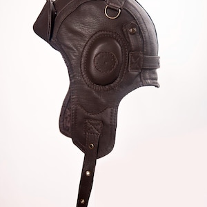 Può includere: Un casco da aviatore in pelle marrone con paraorecchie, sottogola e una piccola visiera. Il casco ha un'estetica vintage, con cuciture visibili e hardware in metallo. Il design ricorda gli equipaggiamenti aeronautici.