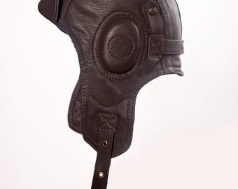 Puede incluir: Casco de aviador de cuero marrón con orejeras, correa para la barbilla y una pequeña visera. El casco tiene una estética vintage, con costuras visibles y herrajes metálicos. El diseño recuerda al equipo de aviación antiguo.