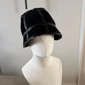 Op de afbeelding: Een zwarte bucket hat met een suède-achtige textuur en leren afwerking. De hoed wordt getoond op een witte mannequinhoofd. De hoed heeft een gestructureerd ontwerp met een licht gebogen rand en een decoratieve leren band.