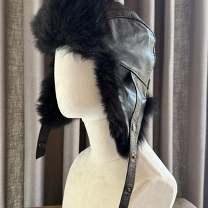 Pode incluir: Um chapéu de caçador em couro preto com forro e acabamento em pelo preto espesso e felpudo. O chapéu inclui abas para as orelhas que se estendem para baixo, presas por tiras finas de couro com botões de pressão metálicos visíveis.