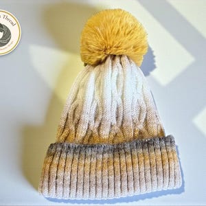 Op de afbeelding: Een gebreide muts met een kabelpatroon en een grote, pluizige pompon. De muts heeft een grijze rand, die overgaat in crème en bruin, en is afgewerkt met een goudgele pompon. Het Warmth & Thread logo is zichtbaar.