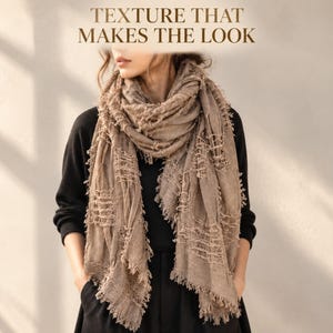 Puede incluir: Una bufanda de color beige con una textura de tejido abierto y bordes deshilachados, sobre una prenda negra de manga larga. El texto "TEXTURE THAT MAKES THE LOOK" aparece impreso en marrón en la parte superior central.