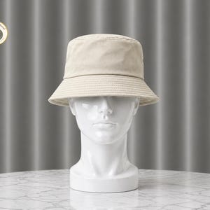 Op de afbeelding: Een beige bucket hat met een gestructureerde rand wordt getoond op een witte mannequinhoofd. De hoed heeft een eenvoudig, klassiek ontwerp en is gemaakt van een zachte, neutrale stof. De hoed is een veelzijdig accessoire.