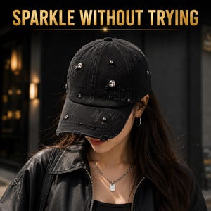 May include: A black distressed denim baseball cap featuring scattered clear rhinestones and the text SPARKLE WITHOUT TRYING printed in gold at the top. The cap has a structured crown, a curved brim, and visible frayed edges on the fabric.