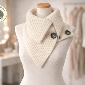 May include: A cream-coloured knitted cowl neck warmer with two dark buttons, displayed on a mannequin. The Warmth & Thread logo is visible in the upper left corner. The background is blurred, with clothing hanging on a rail.