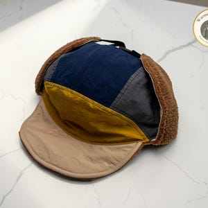 May include: A multi-colored trapper hat with a brown faux fur lining and ear flaps. The hat features a navy blue, gray, and yellow patchwork design on the crown and a tan brim. The hat is designed to keep the wearer warm in cold weather.