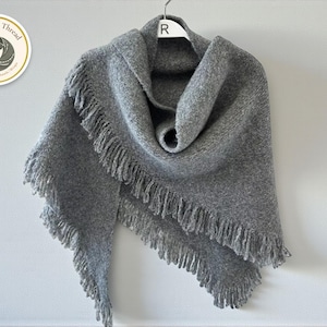 Peut inclure: Un châle gris avec des franges, suspendu à un cintre blanc. Le châle est fait d'une matière douce et texturée. Le logo sur la gauche indique "Warmth & Thread".