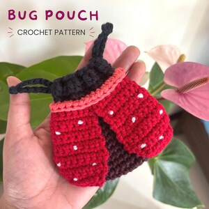 Könnte beinhalten: Ein gehäkeltes Täschchen in Marienkäferform mit roten Flügeln mit weißen Punkten, einem schwarzen Kopf und schwarzen Fühlern. Das Täschchen hat einen rosa Rand und einen Kordelzug. Der Text BUG POUCH CROCHET PATTERN ist oben zu sehen.