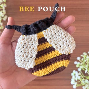 May include: A crocheted bee pouch featuring horizontal yellow and dark brown stripes on the body, two cream-colored wings, and a dark brown top section with two small antennae. The text BEE POUCH is printed in yellow at the top of the image.