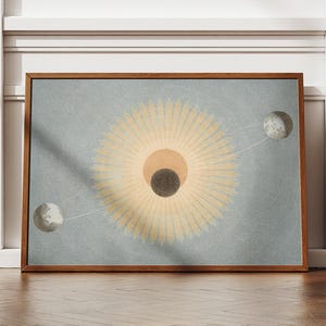 Può includere: Una stampa artistica incorniciata con un design celeste. L'opera raffigura una forma centrale simile al sole con linee radianti, un cerchio scuro e due oggetti simili alla luna in orbita. La tavolozza dei colori include tonalità beige, grigie e marroni.