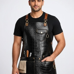 Puede incluir: Delantal de cuero negro con textura de piel de cocodrilo, correas de cuero marrón y múltiples bolsillos. El delantal tiene un bolsillo para herramientas y una toalla. El delantal lo lleva una persona que viste una camiseta negra.