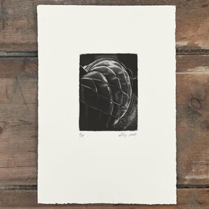 May include: A black and white print of a detailed, textured object, possibly a fruit or a decorative item, with a grid-like pattern. The print is on a white paper with the text "2/5" and "w.ley 2025".