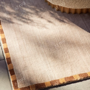 May include: A rectangular beige rug with a brown and tan checkered border. The rug is partially rolled up, revealing a textured underside. The rug is placed on a gray tiled surface, with shadows cast across it.
