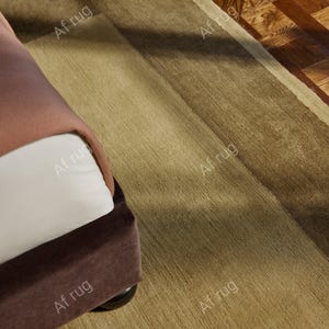 May include: A rectangular area rug in shades of tan and brown, with a darker border. The rug is placed on a wooden floor, and a portion of a brown upholstered piece of furniture is visible on the left side. The rug has the text "Af rug" repeated on it.