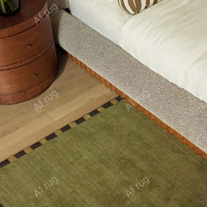May include: A bedroom scene featuring a green rug with a black and gold border, a bed with a patterned headboard and a brown and white striped bolster pillow. A wooden nightstand with three drawers is also visible.