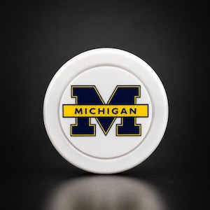 May include: A circular white plastic disc featuring a dark blue block letter M with a yellow horizontal bar across the center containing the word MICHIGAN in dark blue capital letters. The design is centered on the smooth white surface.