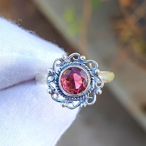 Puede incluir: Un anillo de plata con una piedra preciosa redonda, facetada y de color rojo intenso engastada en un bisel. El engaste está rodeado por un borde decorativo de cuerda trenzada y un marco exterior de bucles de alambre de plata con cuentas.
