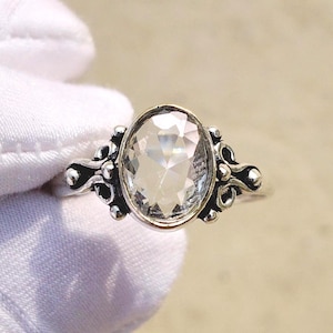 Op de afbeelding: Een zilveren ring met een heldere, ovale edelsteen. De edelsteen is gezet in een decoratieve zilveren band met sierlijke details aan weerszijden. De ring wordt getoond tegen een effen achtergrond.