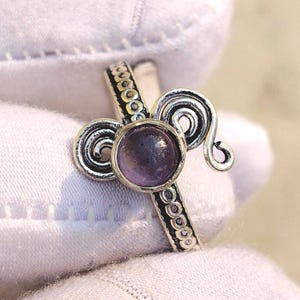 Puede incluir: Un anillo de plata con una piedra preciosa redonda y púrpura en el centro. El anillo presenta remolinos decorativos a ambos lados de la piedra preciosa y una banda detallada. El anillo está sostenido por una mano enguantada.