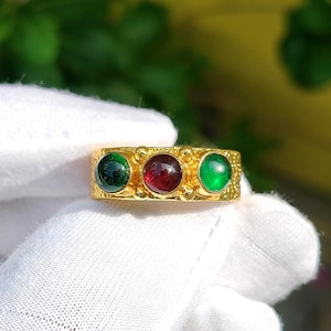 Op de afbeelding: Een goudkleurige ring met een textuurrijk, korrelig oppervlak en drie ronde, cabochon geslepen edelstenen in een horizontale rij. De middelste steen is dieprood, geflankeerd door twee levendige groene stenen aan weerszijden in gouden zettingen.