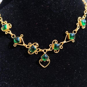 Può includere: Una collana dorata con pietre preziose verdi. La collana presenta pietre quadrate e a forma di diamante incastonate in una montatura dorata decorativa. Il design include volute e una catena.