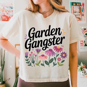 May include: Cream-colored t-shirt with the words "Garden Gangster" in black script above a floral design. The flowers are in shades of pink, purple, and yellow, with green leaves. The shirt is a casual, comfortable style.
