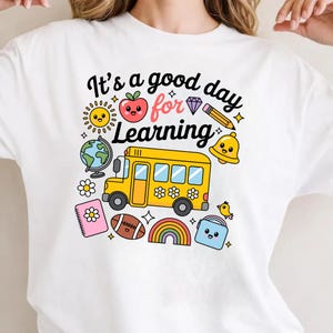 May include: White t-shirt with a colorful graphic design featuring a yellow school bus, a smiling sun, an apple, a globe, and the text "It's a good day for Learning". Other elements include a rainbow, a football, and a bell.