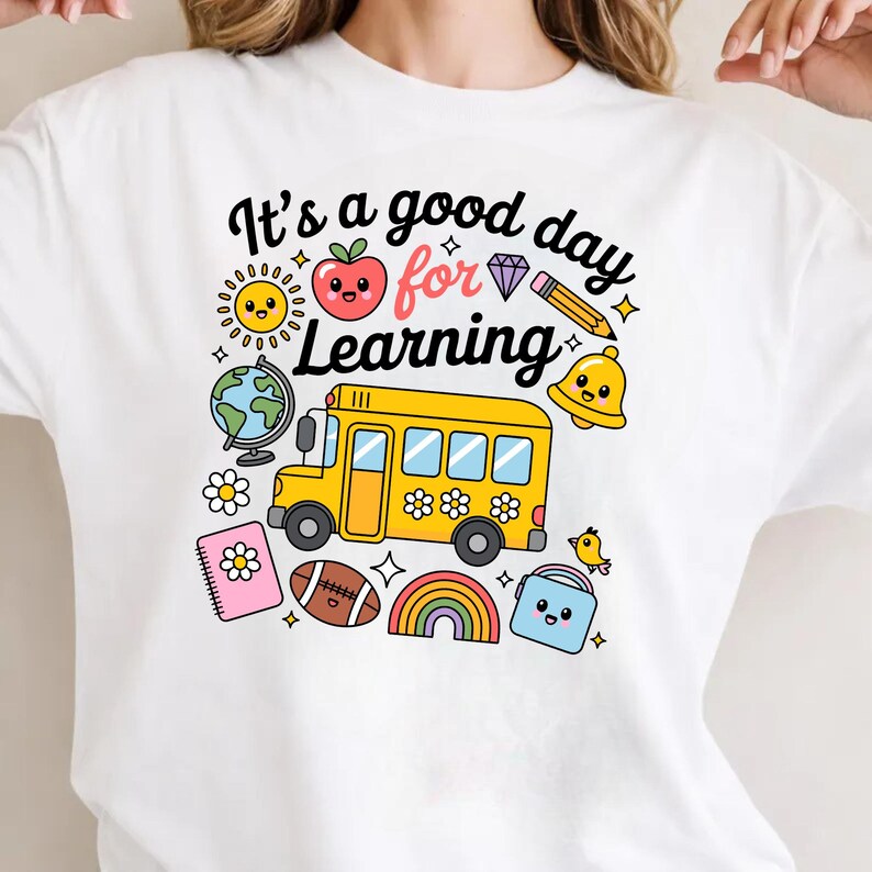 May include: White t-shirt with a colorful graphic design featuring a yellow school bus, a smiling sun, an apple, a globe, and the text "It's a good day for Learning". Other elements include a rainbow, a football, and a bell.