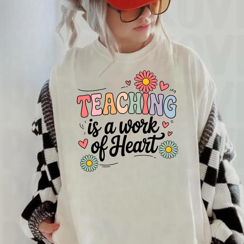 May include: Cream-colored t-shirt with the text "TEACHING is a work of Heart" in colorful, playful lettering. The design includes daisy flowers and hearts. The shirt is a casual, everyday wear item.