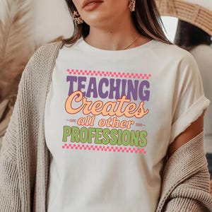May include: White t-shirt with the text "TEACHING Creates all other PROFESSIONS" in purple, orange, and green. The text is framed by pink and purple checkered patterns. The shirt is a casual wear item.