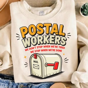 May include: A cream-colored crewneck sweatshirt featuring a graphic of a mailbox with a red flag. The text reads POSTAL WORKERS WE DON'T STOP WHEN WE'RE TIRED WE STOP WHEN WE'RE DONE in yellow, black, and red lettering on the front of the garment.